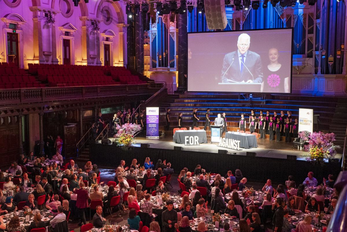 Wide view of the audience and the stage. Tony Jones is on the stage with the choir.