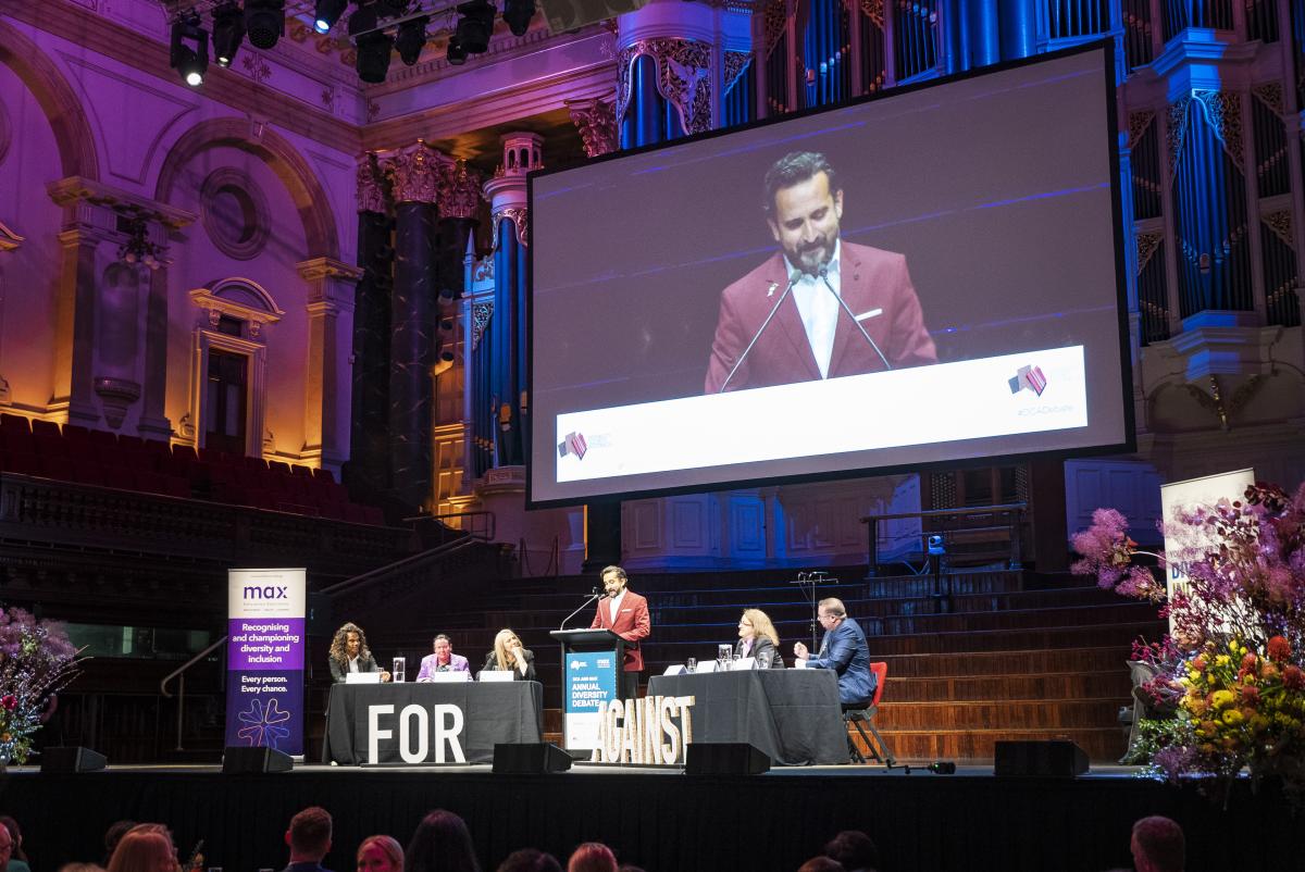 Image of the stage, the panel are seated with Sami Shah speaking at the lectern and his image on the screen above him.