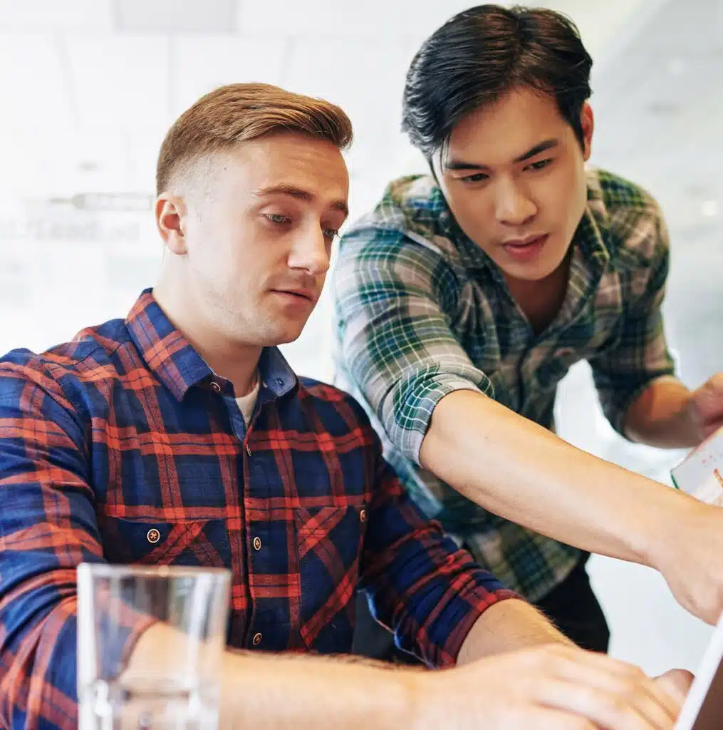 One young white man sitting in front of a lap top with one young Asian man standing next to him pointing at the laptop screen. They both have short hair and plaid shirts with the sleeves folded up to their elbows.