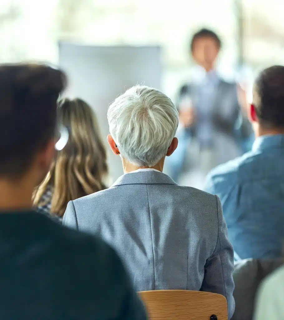 Person standing and talking in the background, in the foreground the backs of people sitting down and listening with focus on a woman with short, blonde or grey hair, an earring and a light blue suit.