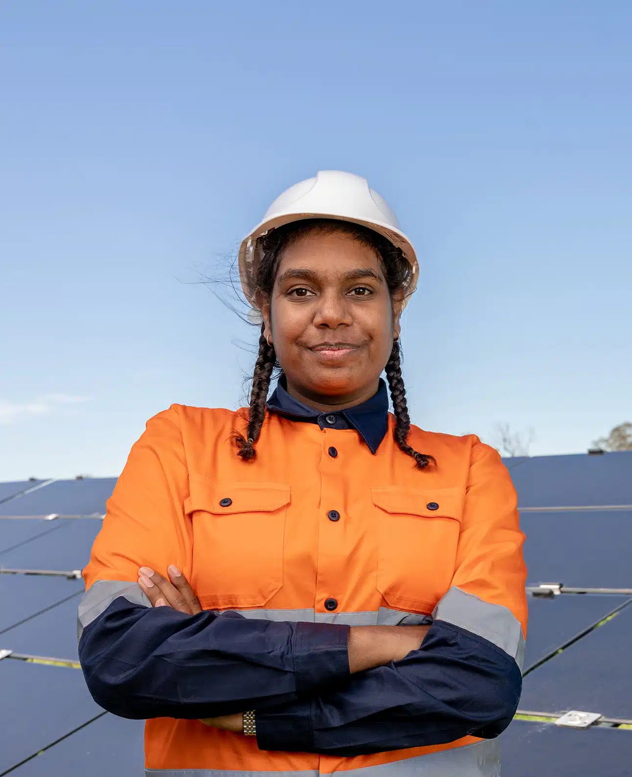 Young First Nations Australian in hi-visibility clothing and safety helmet, smiling with her arms crossed, in front of a solar farm