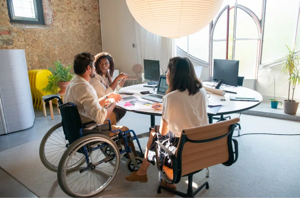 Three workers around a table in an office setting. One is a woman with brown skin who appears to be speaking with a man with dark hair and light skin, sitting in a wheel chair. The third person is a woman with dark her, her face away from the camera, listening to the conversation. The setting is bright and airy.