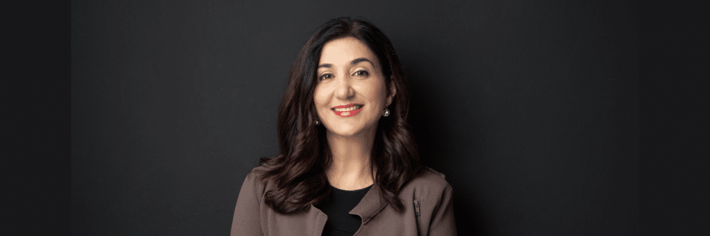 Lisa Annese, a women with long, dark brown hair, a brown suit, red lipstick, smiling at the camera in front of a dark grey background