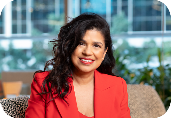 Headshot of Sunita Gloster. She has long, curly, dark hair, brown skin and is wearing an orange business suit and is smiling at the camera.
