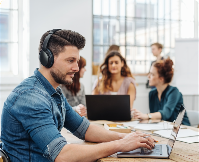 Person with headphones working on computer at desk with group of people working behind them