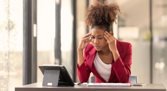 Woman in red suit sits alone at a desk with her eyes closed and her hands on her temples.