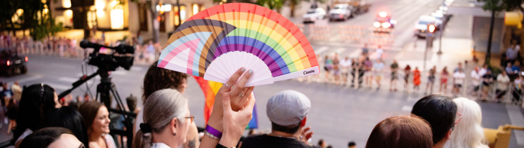 Fan with Progress Pride Flag being held by two arms with blurred background of street waiting for parade