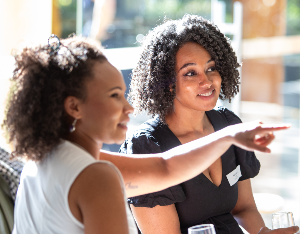 Two women having a conversation. One is pointing with her left hand while the other is smiling.