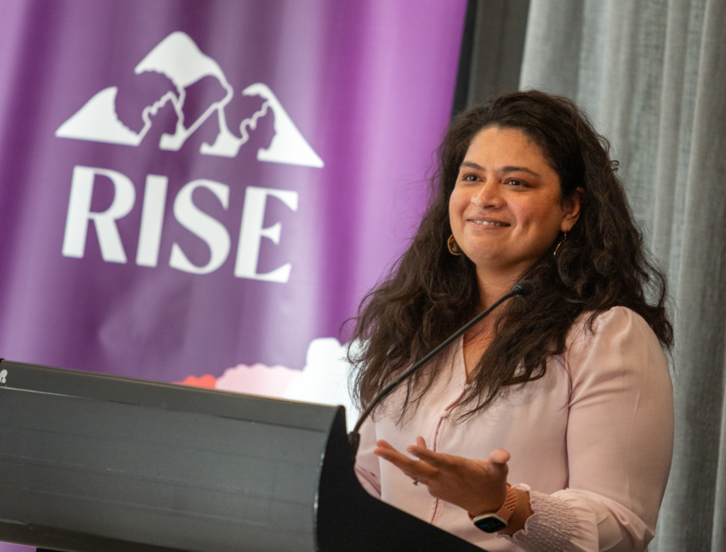 Women speaking at a podium in front of a RISE banner.