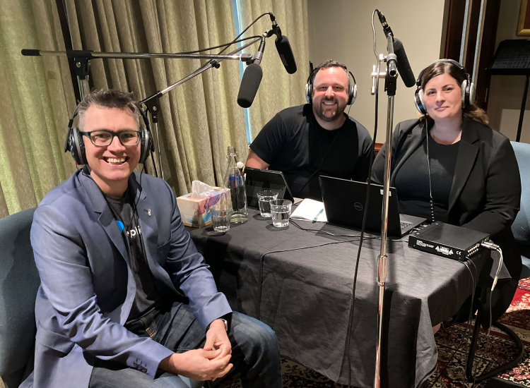 Three individuals sitting around a podcasting table smiling and looking at the camera. There is a microphone in front of each person