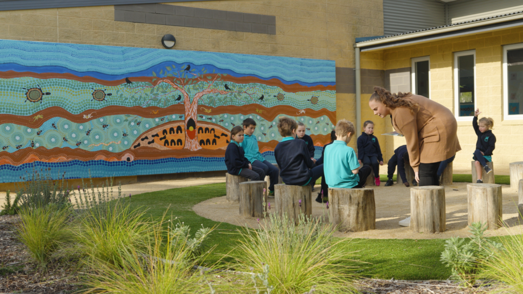 School children sit outside in front of a mural with First Nations artwork.