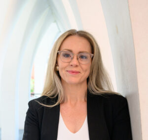 A photo pf Grace Molloy, a woman with long, straight blonde hair wearing clear framed glasses and a navy blazer over a white blouse.